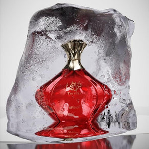 A red glass perfume bottle with a metallic gold neck, set inside a large clear ice sculpture on a reflective surface against a light gray background.